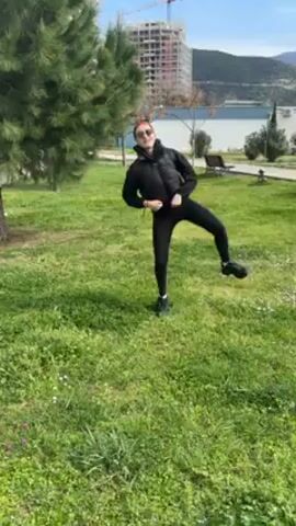 A woman in a black vest, sunglasses, and leggings is outside doing stretching exercises on a grassy area on a sunny day. She is facing the camera and moving her right leg in front and to the side. There are trees and a building in the background.