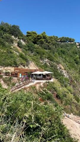 The video opens with a panoramic view of a steep, green, tree-covered cliff face on a bright, sunny day. A clear blue sky is visible above. A restaurant with white umbrellas and tables is perched on a ledge halfway up the cliff, with people seated at the tables. A wooden walkway with railings leads up to the restaurant from the left. Further down the cliff and to the right, a beautiful white-pebble beach is lined with rows of thatched umbrellas and lounge chairs. The turquoise water of the sea is calm and clear, with several people swimming and enjoying the beach. The camera pans slightly to the right, revealing more of the beach and the coastline, with some buildings visible on the clifftop in the distance. The overall impression is one of a picturesque and relaxing vacation destination.