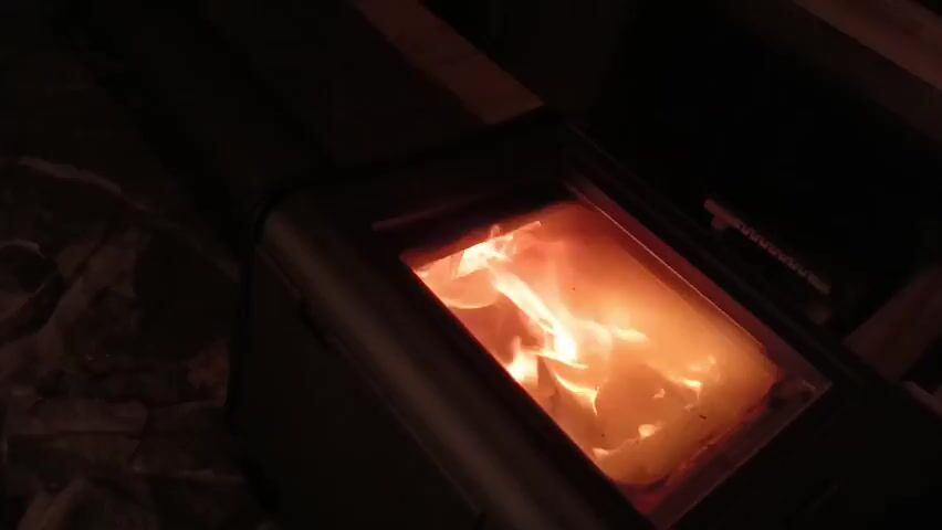 A wood-burning stove glows in the dark, the bright orange flames visible through the glass door. The stove itself is dark in color, likely black or dark gray. The surrounding area is dimly lit, showing a wood floor or base on one side and what appears to be stone or brick on the other. The camera angle is slightly high, looking down at the stove. There is no visible movement other than the flickering of the flames within the stove.
