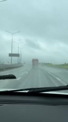 The video shows a drive through a wet, overcast landscape from inside a car.

The first shot (0:00-0:01) is from the driver's perspective, looking out a rain-streaked windshield onto a wet highway. A large red truck with a white "K" logo on its back is visible ahead, followed by other vehicles. The sky is grey and heavily clouded. Streetlights line the left side of the road.

The second shot (0:01-0:02) briefly shows the car's interior, specifically the right side of the dashboard, a speaker grille, and the wet front side window.

The final shot (0:02-0:04) pans to the right, showing the view from the car's side window. A lush green embankment covered in small yellow flowers slopes upwards, topped by a dense line of evergreen trees against the grey sky. The wet road surface is visible in the foreground.

The scene depicts a rainy daytime drive on a highway in what appears to be a rural or semi-rural area near Kukushtan, Russia, characterized by green hills and forests.