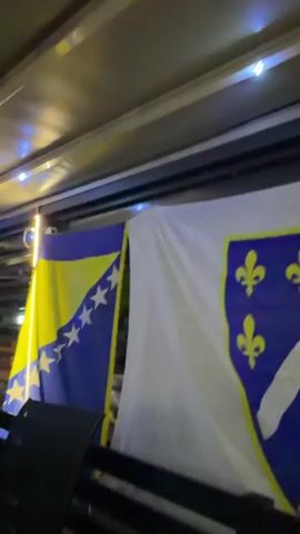 The video begins with a close-up view of flags, including the Bosnian flag with its blue and yellow colors and stars, and another flag with a fleur-de-lis emblem. The camera then pans across a room with seating and a television screen. The television displays a soccer match, with two players in uniform visible on screen. One player is wearing a blue jersey with the number 4, and the other is in a white jersey with red and black stripes. Other people are visible in the background of the televised game. The room appears to be an indoor space with ambient lighting, possibly a lounge or viewing area.