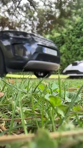 A robotic lawnmower is visible in the foreground, partially obscured by grass. The lawnmower is white with yellow accents and has black wheels. In the background, a dark-colored SUV is parked. The scene appears to be a lawn with trees and bushes. The lighting suggests it is daytime.
