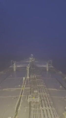 A drone footage taken from behind an oil tanker shows its deck. The sky is dark, and the sea is deep blue and somewhat murky. The tanker's deck is shiny and reflects the light. The drone moves forward, capturing a central view of the deck, highlighting the pipes and machinery, with the bow of the ship visible in the distance. The overall impression is that of a vast, empty ocean and the solitary presence of the tanker.