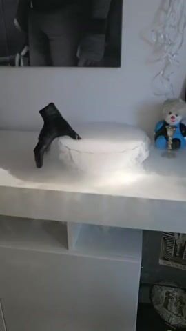 A black gloved hand with a red painted fingernail rests on the edge of a white ceramic bowl. The bowl is filled with a white mist, creating a dramatic effect. The mist billows over the sides of the bowl and onto the white surface it rests on. A clear glass pitcher is shown being tilted over the bowl, with liquid appearing to pour from it into the mist. The background shows a portion of a black and white framed picture hanging on a light-colored wall. The scene appears to be set on a white shelf or counter.