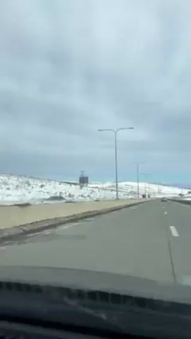 A car drives along a straight highway on a cloudy day. The road is gray with white lane markers, flanked by snow-covered terrain. Tall streetlights line the roadside. A vehicle is visible in the distance, further down the highway. The sky is a muted blue-gray, suggesting a cool, overcast climate. The view is captured from inside the moving car, focusing on the road ahead and the surrounding landscape.