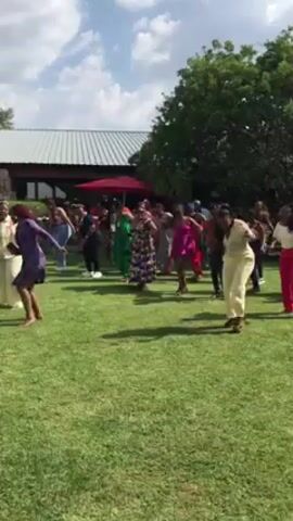 A group of women are dancing on a grassy lawn, seemingly having fun, under a partly cloudy sky. Some of the women are wearing brightly colored outfits. In the background, there is a building with a metal roof and a red umbrella. A man in a white shirt passes in front of the camera, obscuring the view for a moment. The overall setting appears to be an outdoor event or gathering.