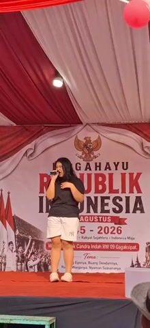A young woman with dark hair is standing on a stage, holding a microphone and singing. She is wearing a black t-shirt and white shorts. Behind her is a large banner with Indonesian text and symbols, including the Garuda Pancasila. The stage is decorated with red and white fabric, and there are Indonesian flags on the left side. The event appears to be a celebration of Indonesia's Independence Day.