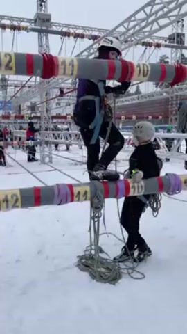 The video shows an outdoor obstacle course in a snowy environment. Several people wearing helmets and safety harnesses are navigating the course, which consists of horizontal bars numbered 10 through 15, connected by ropes and cables. The participants are walking or climbing across these bars. A few people on the ground appear to be assisting or observing. The sky is overcast, and the ground is covered in snow. The course is constructed of metal scaffolding, and there are stadium seats visible in the background.