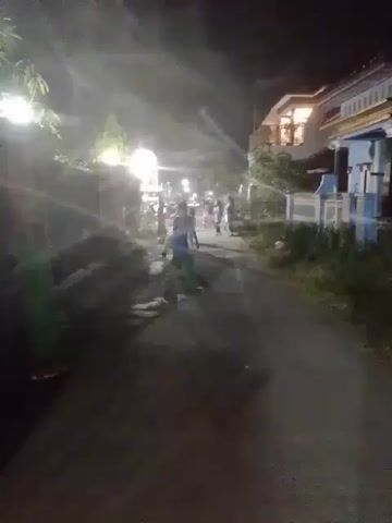 At night, the video shows a group of children playing on a narrow road lined with houses. The road is illuminated by streetlights, which cast long shadows and create a hazy atmosphere. The children are scattered along the road, some walking and others running. One child stands to the side, observing the group. The houses on either side of the road are visible, with some showing lit windows. The overall scene suggests a casual, neighborhood gathering at night.