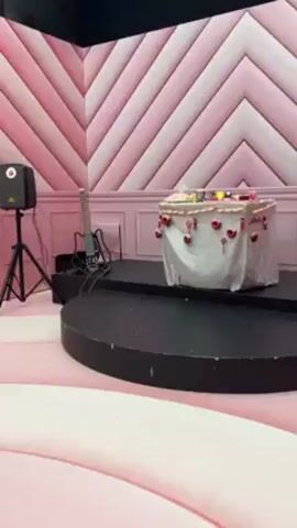 The video shows a stage in a room with a pastel pink chevron patterned wall. A black, rounded stage sits in the center, with a smaller, rectangular platform elevated slightly on top. On this platform is a small table covered with a white cloth decorated with dangling red heart ornaments. Various items are set on the table’s surface. To the left of the stage, a guitar and a speaker on a stand are visible. The room is likely a set or a themed event space, given the decorative elements and the stage setup. Music plays in the background.