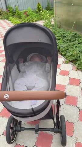 A baby in a gray onesie and white socks sleeps in a stroller outdoors. The stroller has a brown handle and is on a tiled patio. Green plants and a fence are visible in the background.