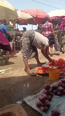 The video shows a woman in a market selecting tomatoes. She wears a beige shirt and a long patterned skirt. She is bending over a display of tomatoes, sorting through them with her hands. A yellow bucket sits nearby. Several other people are walking around in the background, and various stalls are visible under umbrellas. Some red onions are in the foreground. A parked motorcycle and a wheelbarrow also feature in the background. The ground is dusty.