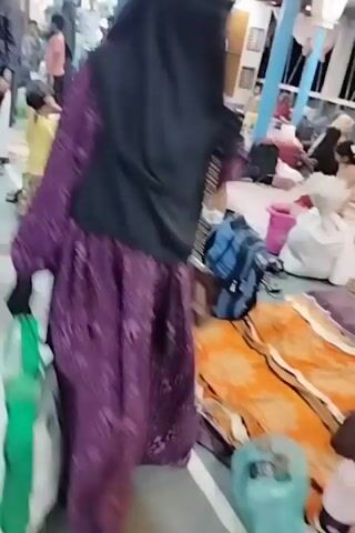 The video depicts a crowded indoor space, likely a waiting hall or transit terminal in Bhola, Bangladesh. Numerous people, including men, women (some wearing hijabs or niqabs), and children, are present. The area appears to be a large, open room with a concrete or tiled floor and overhead fluorescent lighting, along with ceiling fans. Blue metal poles are visible, possibly delineating sections of the space.

Many individuals are carrying luggage, including suitcases and backpacks, indicating travel or relocation. Some men are seen carrying large bags on their shoulders or heads as they navigate through the crowd. Other people are seated on the floor amidst their belongings, suggesting a period of waiting or rest. The atmosphere is bustling and congested, with people moving in various directions or settling down. The general attire is casual, typical of South Asian regions. No specific time of day is discernible, but the lighting suggests it's either daytime or a well-lit interior. No discernible text is visible in the video.