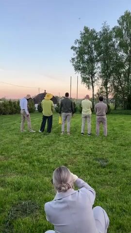 A video opens with a shot of a person with blonde hair pulled into a bun crouched on a lawn, filming five men standing on the grass facing away from her. They are all wearing somewhat mismatched outfits of muted colors. One is holding a yellow grocery bag.  A man moves toward the front and picks up a yellow object, then performs a dance move with it in front of his crotch. A second camera operator enters from the left side of the screen.