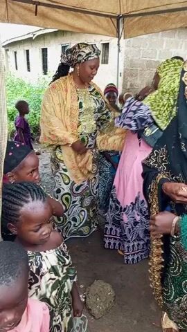 Here's a concise description of the video:

The video shows a group of African women and children standing under a tent-like structure. The women are dressed in colorful, traditional patterned clothing and head coverings. They are smiling and talking amongst themselves. Some of the children are also looking at the women and interacting with them. The background shows a building and some greenery. The overall atmosphere appears to be festive or celebratory.