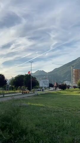 The video pans right across a grassy field towards a road with trees and buildings beyond it. There are several cars driving on the road, and a few people walking. In the background, there are mountains. A large building, possibly under construction, is visible on the right side of the frame. The sky is blue with some clouds and a contrail. The lighting suggests it is late afternoon or early evening.