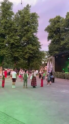 The video captures an outdoor event with a stage set up on the right side.  Several people are present, with many gathered in a loosely organized circle in the foreground, seemingly participating in or observing a dance.  A stage with lighting and two performers, one playing an accordion and the other singing, is visible on the right.  Tall trees with lush green foliage frame the scene, and string lights are visible overhead. The sky shows a soft gradient of pink and purple, suggesting it is either early morning or late evening.  The ground is a paved or concrete surface.  Several individuals are dressed in what appears to be traditional or folk-inspired clothing, particularly a woman in a plaid skirt and a man in a vest.  The overall atmosphere appears festive and community-oriented.