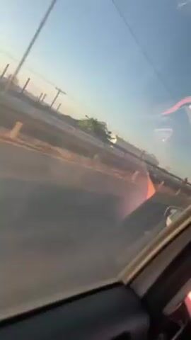 The video, shot from the inside of a moving car, shows road construction workers wearing orange protective gear and safety helmets on a highway. Some of them are seen moving a traffic cone. The weather appears sunny, with a clear blue sky.  A building and power lines are visible in the background. The car continues to move along the highway.