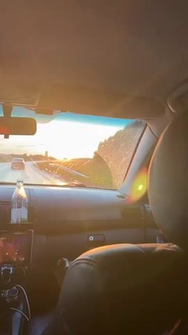 The video shows the interior of a car driving on a road during sunset. The camera is positioned behind the driver and passenger, looking out through the windshield. The sun is visible in the distance, casting a warm glow on the scene. The dashboard of the car is visible, with a GPS device and a bottle of water mounted on it. The driver's hands are on the steering wheel, and the passenger's feet are visible. The car is moving along a paved road, with trees and fields on either side. The windshield wipers are visible, and it appears that some water is being sprayed onto the windshield, creating streaks that refract the sunlight. The overall impression is one of a peaceful and scenic drive.