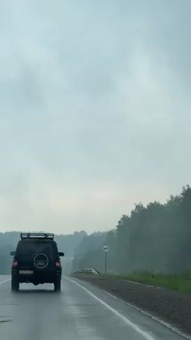 A black SUV is driving on a wet road. The sky is cloudy and overcast. The road is bordered by trees on the right side of the image. There is a road sign on the right side of the road. The SUV has a spare tire mounted on the back and a roof rack. The video appears to be recorded from another vehicle driving behind the SUV.