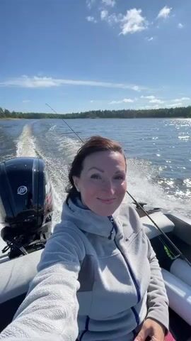 A woman with dark hair is taking a selfie while sitting in a boat. The boat is moving and creating a wake. The woman is wearing a gray hooded jacket. There is a black outboard motor on the back of the boat. The sun is shining and the sky is blue with some clouds. There are trees on the shore in the distance.