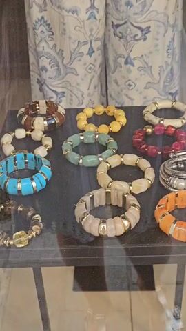 The video shows a close-up view of a display of bracelets behind glass. The bracelets are made of various beads and materials, including wood, stone, and metal. They are arranged on a dark surface. In the background, a person's arm is visible, and a white and blue patterned fabric is hanging. The reflection in the glass shows a street scene with a white car and buildings.