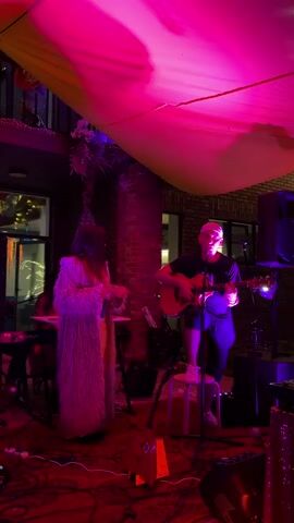 The video shows a live music performance taking place outdoors at night. The scene is illuminated by pink and yellow lighting. A woman wearing a long, fluffy coat and a yellow dress is standing on the left, holding a microphone and singing. She has long, dark hair. To her right, a man wearing a black t-shirt, dark pants, and a baseball cap is sitting on a stool and playing an acoustic guitar. He is facing the woman and singing as well. In the background, there is a brick wall, some plants, and musical equipment including keyboards and speakers. A yellow tarp is stretched overhead, creating a canopy. The floor is covered with a red, patterned rug. There are other people visible in the background, suggesting an audience or other performers. The overall atmosphere appears to be intimate and perhaps a bit bohemian.