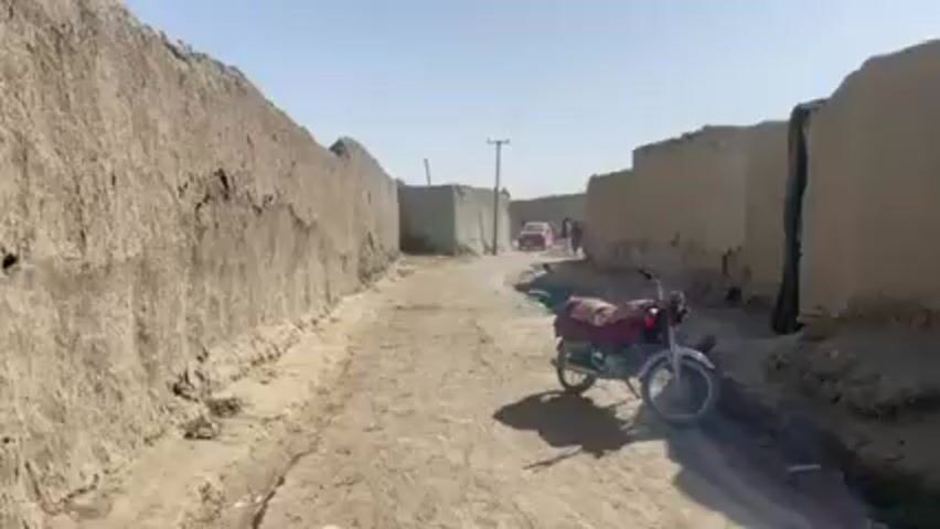 A narrow dirt road runs between tall, weathered mud walls, leading toward a distant red car and a small group of people. A motorcycle with a red and brown patterned cover sits parked on the right side of the road. A power pole stands near the center of the frame. A thin, shallow stream of water runs along the road. The sky is clear and bright. The overall setting appears to be a rural or underdeveloped area.