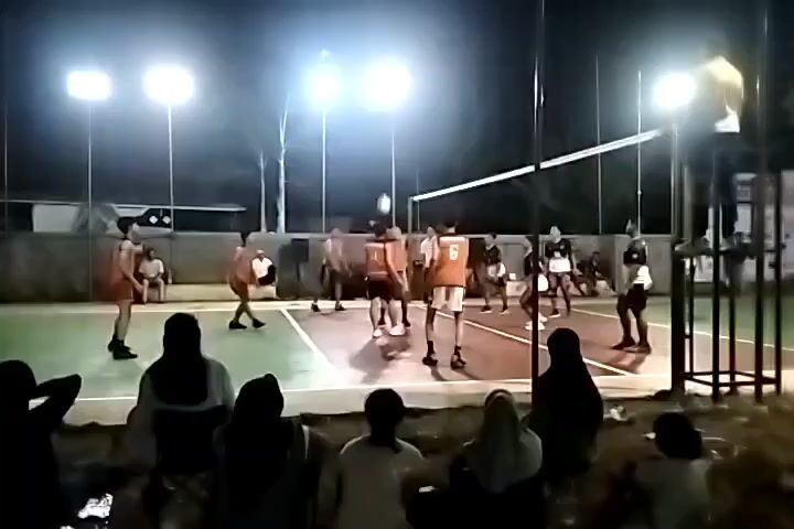A volleyball game is being played at night on an outdoor court. The court is lit by several bright floodlights. There are players on both sides of the net, and spectators are watching from the sidelines. The players are wearing athletic attire. One player is in the process of serving the ball, and another player is jumping to hit it. The spectators are seated on the ground and appear to be casually observing the game. The environment is dark, with the floodlights illuminating the court and the players.