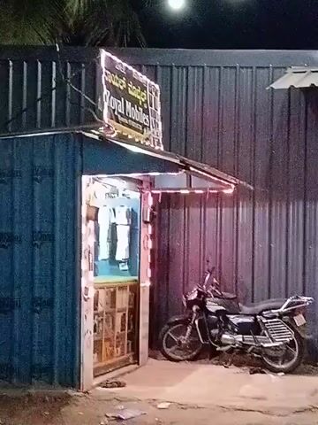 A small shop, possibly a mobile phone repair or sales business, is situated within a blue corrugated metal structure. The shop is illuminated by strings of colorful LED lights, primarily white and pink, outlining the entrance and the sign above it. The sign, also lit, displays text in what appears to be a South Indian language, along with "Royal Mobiles." Inside the shop, displays of mobile phones or accessories are visible. To the right of the shop, a black motorcycle is parked. The overall environment suggests an outdoor setting at night, with some foliage visible against a dark sky. The scene is static, with no visible human activity or interaction.