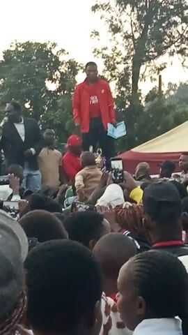 A man in a red shirt and black pants stands before a crowd of people. He holds a booklet in his left hand and gestures with his right. He appears to be speaking to the crowd, which is seated on the ground. The scene is outdoors, with trees visible in the background. A large tent or canopy is erected behind the man. The sky is visible, suggesting it is either early morning or late afternoon. The people in the crowd are mostly looking towards the man, with some holding cell phones. The orientation of the video is rotated 90 degrees counter-clockwise.