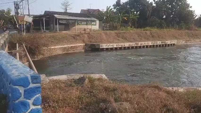 The video shows a canal with flowing water. On the left side of the canal, there is a bridge railing with blue and white stone decorations. Beyond the bridge, there are buildings and vegetation. The canal itself is lined with concrete and stone walls. The water appears to be somewhat murky, with ripples and small waves indicating movement. In the distance, there are trees and a clear sky. The overall scene suggests a rural or suburban environment, possibly in a tropical or subtropical region given the vegetation.