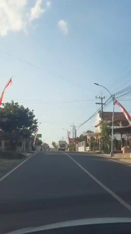 A car's perspective shows a two-lane road with white lane markings. A motorbike with two people is traveling away from the camera. To the right, a white truck is also traveling away from the camera. Along the sides of the road are houses and trees. Red and white flags are visible on poles. The sky is clear with a few clouds.
