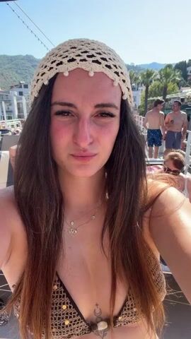 A woman with long dark hair and a crocheted hat adorned with pearls takes a selfie on a boat. She is wearing a sequined bikini top. Behind her, people are visible on the deck of the boat, some wearing swimwear. In the background, there are buildings on a hillside and a large ship. The scene appears to be a sunny day, possibly in a coastal town.