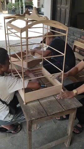 Three people are working on a wooden birdcage. The birdcage is tall and intricate, made of thin wooden slats. It has a rectangular base, a series of horizontal bars, and an arched top with decorative carvings. The cage is sitting on a rough wooden table. Two men are actively working on the cage. One man, wearing a light-colored t-shirt and dark pants, is kneeling and using a tool to attach a piece of wood inside the cage. His body is partially inside the cage. Another man, wearing a dark t-shirt and a cap, is also kneeling and working on the cage, looking at it from the opposite side. His hands are also inside the cage. A third person's hands are visible on the right side, resting on the table or the cage. The environment appears to be outdoors, possibly a workshop or a backyard, with a building in the background and some motorcycles parked in the distance. There are sawdust particles on the table.
