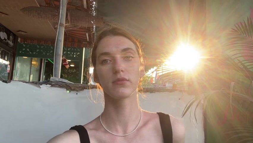 A fair-skinned person with light brown hair pulled back, wearing a black tank top and a pearl necklace, looks directly at the camera. The person is outdoors with sunlight shining directly behind them, creating a bright glare. Behind the person is a white wall with a tropical plant painted in pink and green. Part of a building is visible in the background to the left, with hanging decorations above. The person remains still throughout the video.
