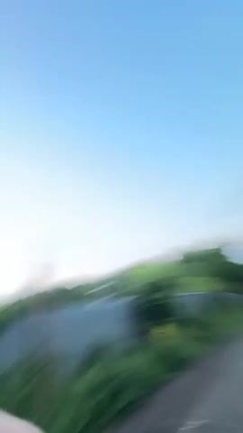 This is a first-person perspective video of a young woman with brown hair, wearing a black hoodie and sunglasses, as she rides on a skateboard. She is outdoors on a paved path, with green foliage and a brown fence lining the path. She smiles and touches her forehead as she looks at the camera. The video is blurred due to movement.