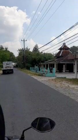 A first-person perspective video shows a paved road with a motorcycle in the foreground. A white truck with text on its side is driving away from the camera on the left side of the road. On the right side of the road, there is a building with a domed roof, possibly a mosque, with a light blue fence in front of it. Trees and power lines are visible throughout the scene. The sky is partly cloudy.