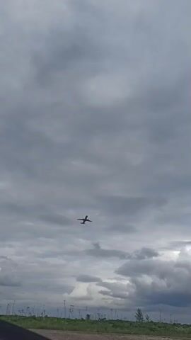The video shows an airplane flying overhead against a cloudy sky. The airplane is dark in color, but with red elements, and the sky is gray. The plane approaches closer to the camera before flying out of view. In the bottom of the screen is a strip of green vegetation.