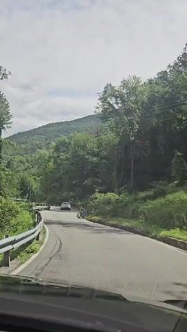 A car is driving on a winding road through a lush green mountainous landscape under a partly cloudy sky. The road has guardrails on the left side, and trees and vegetation line both sides of the road. There are a couple of other cars visible further ahead on the road. The sun is shining, casting shadows on the road.