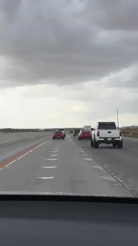 The video shows a view from inside a vehicle traveling on a highway. Several other vehicles are visible on the road ahead, including red cars and a white pickup truck. The sky is overcast with dark clouds. The road appears to be asphalt and has lane markings. The scene suggests a typical highway driving experience under cloudy weather conditions.