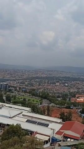 The video showcases a broad aerial view of a city spread across a hilly landscape. The sky overhead is overcast with dense clouds. In the foreground, there are rooftops of several buildings, one notable for its solar panels, and a red roof. A tall building stands on the left side. The cityscape in the distance is characterized by dense, mostly low-rise buildings interspersed with patches of greenery and trees. The landscape gently rises towards the horizon, where faint hills or mountains are visible. The overall tone is muted, owing to the overcast weather.