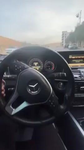 The video shows the interior of a car, likely a Mercedes-Benz, while it's being driven. The initial view is of the steering wheel and dashboard, showing the speedometer which reads around 37 km/h. The temperature is indicated as 11.5 degrees Celsius. The view then shifts to the rearview mirror, reflecting the driver's face. The dashboard, infotainment system, and car interior are visible. The exterior scene appears to show a road in a suburban area with buildings in the background.