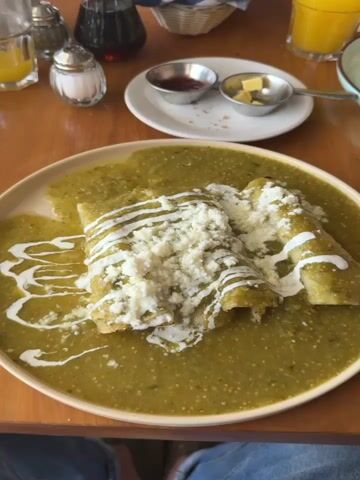 The video shows a breakfast meal at a restaurant. The first scene focuses on a plate of enchiladas covered in green sauce, queso fresco, and crema, sitting on a wooden table. The camera then pans to the right, revealing a plate of pancakes topped with yellow sauce, powdered sugar, and raspberries. A glass of orange juice sits nearby. The camera pans further to show more of the table setting, including a napkin-wrapped fork, silverware, a small plate with butter and jam, and a sugar and syrup dispenser. Finally, the camera zooms out to give a view of the restaurant's interior. There are multiple people seated at tables. The overall atmosphere appears lively and casual.