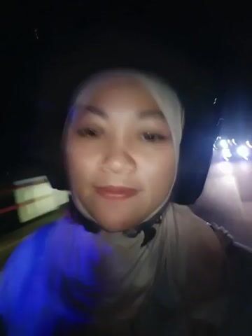 A woman wearing a light-colored hijab and a dark helmet is riding on a motorcycle at night. She is looking directly at the camera and smiling. The background is dark, with some blurred lights suggesting a road or street scene. The video appears to be a selfie taken from the perspective of someone riding behind her or holding the camera.