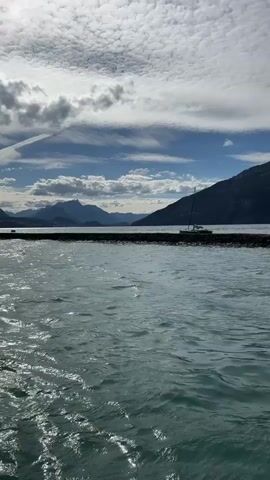 The video is a panoramic shot of a body of water with mountains and trees in the background. The water is a beautiful turquoise color and there are some ripples on the surface. The sky is blue with white clouds. A sailboat is visible on the water in the distance. There is also a stone pier or breakwater in the foreground. The mountains are covered in trees and appear to be quite large. The overall scene is very serene and picturesque.