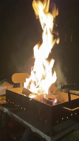 A vertical video captures an outdoor scene at night, featuring a metal grill or brazier containing burning wood. Bright orange flames rise high from the wood, emitting smoke and occasional sparks into the dark background. The metal container, rectangular in shape, has small holes punched along its sides. A lighter-colored, possibly wooden, surface is visible to the left of the container, and a darker, perhaps metallic, object is to its right. The ground or table supporting the container is not clearly visible. The overall lighting is provided by the fire itself, creating a warm glow against the dark surroundings. No people are visible.