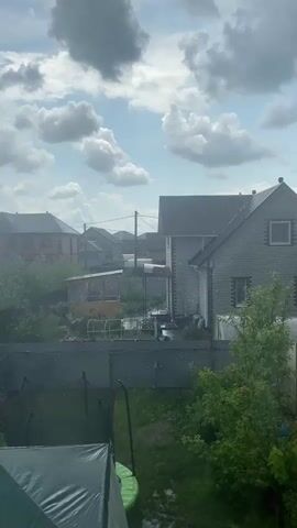 The video shows a view of a backyard with houses in the background. It is raining, with visible raindrops falling. There is a trampoline in the foreground, along with some trees and a fence. The sky is cloudy with some patches of blue. The scene is likely a suburban or rural setting.