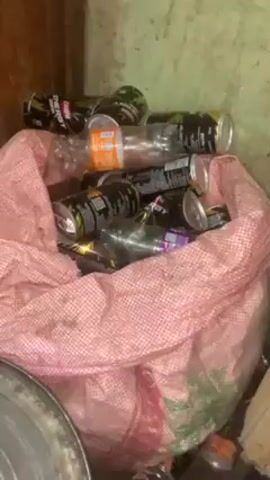 Here is a description of the video:

The video shows a pink checkered bag filled with discarded items.  Visible within the bag are numerous empty cans, primarily of what appear to be energy drinks, along with a few plastic bottles. The bag is situated in what seems to be an enclosed space, possibly a room or storage area.  A metal object, likely a pot or bucket, is visible to the lower left of the bag. The lighting is dim, contributing to a somewhat cluttered and unkempt appearance.
