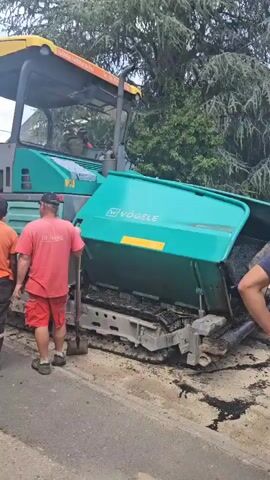 In Hévíz, Hungary, two individuals are observed near a Vögele asphalt paver. One person, wearing a red t-shirt and shorts, stands with their back to the camera, holding a tool. The other individual, in an orange t-shirt and dark pants, faces away, and a third person's legs are visible on the right. The asphalt paver, a large, teal-colored machine with a yellow canopy, is positioned on a road surface that appears to be undergoing construction or repair. Black asphalt is visible on the ground and within the machine's laying mechanism.

The scene suggests an active construction or road maintenance operation. The presence of the asphalt paver, tools, and workers indicates that asphalt is being laid or compacted. The lighting and the presence of green foliage in the background suggest it is daytime, and the weather appears to be overcast or partly cloudy.

Visible text includes "VÖGELE" prominently displayed on the side of the paver. On the back of the red t-shirt, text appears to read "TERRA ÉPÍTŐ HUNGÁRIA". The website "www.uzsonu.hu" is visible on the yellow canopy of the paver. The overall atmosphere seems professional and focused on the task at hand.